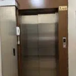 Koyu ahşap çerçeve içine alınmış, kapalı, paslanmaz çelik asansör kapıları merkezi kaplıyor. Sağdaki ahşap yüzeyde "3. KAT" yazılı metal plaka yerini korurken, hemen altındaki kontrol paneli kırmızı dijital "1" rakamını ve yanan yukarı okunu gösteriyor. Asansörün solundaki çelik sütun, üstte havalandırma ünitesini, altında sarmal kablolu dahili telefonu barındırıyor. Genel iç aydınlatma zemindeki mermer döşemeyi aydınlatıyor.