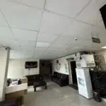 Yapay tavan ışıkları, beyaz kare fayanslarla döşenmiş bekleme salonunu aydınlatıyor. Sol taraftaki ahşap bankonun arkasında, alt kısmına üçgen flamalar asılmış siyah bir televizyon duvarı kaplıyor. Mekanın merkezini siyah dikdörtgen bir sehpa doldururken, sağ arka ve sol duvar diplerini koyu kahverengi deri kanepeler çevreliyor. Sağ ön köşedeki beyaz su sebili ile yanındaki siyah mermer desenli silindir çöp kutusu alanı daraltırken, tavandan sarkan ışıklı "EXIT ÇIKIŞ" tabelası arkadaki karanlık koridora açılan kapıyı işaret ediyor.