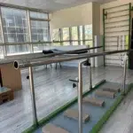 Ön planda, yeşil metal tabanlı ve çelik tırabzanlı paralel yürüme aleti, mavi zeminine aralıklı dizilmiş dikdörtgen ahşap blokları çevreliyor. Sol taraftaki geniş pencerelerden içeri dolan parlak gün ışığı, gri ahşap desenli zemini ve pencere önündeki siyah sandalyeyi aydınlatıyor. Odanın sağ arka duvarını kaplayan yeşil iskeletli tırmanma merdiveni renkli direnç bantlarını taşırken, hemen yanındaki geniş ayna pencereleri ve dışarıdaki binaları yansıtıyor.