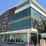 Parlak gün ışığı, sol cephesini kahverengi panellerin, geri kalanını ise yatay beyaz şeritli geniş mavi camların kapladığı köşe binayı aydınlatıyor. Zemin katın cam kapısının ardında mavi ceketli ve koyu renk kasketli yaşlı bir erkek dışarıya bakarken, kaldırımdaki ahşap bank ve kısa erişim rampası binanın girişine yönlendiriyor. Binanın sağ köşesinde dalgalanan Türk bayrağının altında, üzerinde "Özel Büyük Asrın Özel Eğitim ve Rehabilitasyon Merkezi" yazan beyaz tabela ve taş kaideli Atatürk büstü, yeşil bir çam ağacıyla yan yana yükseliyor.