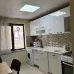 Tavandaki kare aydınlatma panelinden yayılan ışık, krem rengi fayans zeminli mutfağı aydınlatıyor. Sağ duvarı boydan boya kaplayan parlak beyaz üst dolaplar kahverengi mozaik desenli duvara yaslanırken, altlarındaki beyaz tezgâh mikrodalga fırını ve lavaboyu üzerinde taşıyor. Tezgahın alt bölümü sırasıyla çamaşır makinesini ve bulaşık makinesini çerçevelerken, dizilimi en sağdaki beyaz fırınlı ocak tamamlıyor. Sol ön köşedeki açık renkli ahşap masayı koyu bordo sandalyeler sarıyor, hemen arkalarındaki beyaz tül perde ise koyu kahverengi çerçeveli pencereden kalorifer peteğinin üzerine doğru dökülüyor.