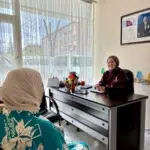 Sol taraftaki tül perdeli geniş pencereden süzülen parlak gün ışığı, ofis odasını aydınlatıyor. Ön planda sırtı dönük oturan, bej başörtülü ve turkuaz üzerine büyük beyaz yaprak desenli elbiseli kadın, ahşap masanın diğer ucundaki kişiye yöneliyor. Masanın ardındaki siyah ofis sandalyesinde oturan yirmili yaşlardaki bordo kıyafetli ve koyu renk desenli başörtülü diğer kadın, sağ elinde tuttuğu kalemiyle evraklara yaslanarak karşısındakine gülümsüyor. Üzerine takvim, kırmızı vazoda renkli çiçekler, isimlik ve siyah bir tabletin dizildiği koyu kahverengi masanın sağındaki bej duvar, ahşap çerçeveli Atatürk portresini sergiliyor.