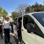 Parlak gün ışığının aydınlattığı parke taşlı kaldırımda, altmışlı yaşlarındaki kır saçlı ve bıyıklı adam, bej rengi kısa kollu tişörtü ve lacivert pantolonuyla sağ taraftaki beyaz minibüsün açık sürgülü kapısına doğru odaklanıyor. Siyah ceketli, koyu renk pantolonlu ve beyaz spor ayakkabılı bir genç, sırtındaki kırmızı ve mavi ağırlıklı desenli çantasıyla minibüsün basamağına tırmanıyor. Adamın sol arkasındaki kaldırımda mavi alışveriş poşeti taşıyan diğer yetişkinler araca doğru yönelirken, arka plandaki yeşil yapraklı ağaçlar ve çok katlı binalar açık mavi gökyüzünün altında sıralanıyor.