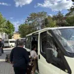 Parlak gün ışığının aydınlattığı parke taşlı sokakta, lacivert yatay çizgili kazak giymiş ellili yaşlardaki kır saçlı adam, sağdaki beyaz minibüsün açık sürgülü kapısından araca binen beş-altı yaşlarındaki iki erkek çocuğuna eliyle destek oluyor. Beyaz uzun kollu tişörtlü ve kahverengi pantolonlu ilk çocuk aracın basamağını tırmanırken, arkasındaki koyu renk şortlu ve yeşil terlikli diğer çocuk da ona tutunarak araca yöneliyor. Aracın içindeki siyah kıyafetli yetişkin erkek çocukların binişini izlerken, adamın solundaki kaldırımda siyah plastik kasalar yükseliyor ve arka plandaki yeşil ağaçların önünde ikinci bir beyaz minibüs park halinde bekliyor.