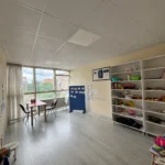 Geniş sol pencerelerden süzülen gün ışığı, gri ahşap desenli zemine sahip boş sınıfı aydınlatıyor. Pencere önündeki küçük kırmızı kare masayı iki siyah sandalye sararken, odanın merkezinde yükselen mavi paravan üzerindeki iğnelenmiş beyaz kağıtları sergiliyor. Sağdaki bej duvarı kaplayan geniş beyaz raflar renkli oyuncakları ve saklama kutularını taşırken, rafların hemen soluna asılı üç bölmeli ahşap çerçeveli portre panosu duvardaki yerini alıyor. Ön planda sağ köşeden uzanan oyuncak davul ve sol köşedeki mavi koni ile küçük tabure odanın sınırlarını çiziyor.