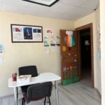 Tavandan sarkan siyah çerçeveli geniş LED lamba, gri ahşap desenli zemine sahip boş sınıfı aydınlatıyor. Ön plandaki metal ayaklı beyaz çalışma masası renkli kalemlerle dolu bir kalemliği ve karton kutuyu üzerinde taşırken, masanın iki yanını siyah sırtlıklı iki sandalye sarıyor. Sol taraftaki bej duvarı, ahşap çerçeveli üç bölmeli Atatürk panosu ile mevsimleri ve ayları gösteren renkli kağıt etkinlikleri kaplıyor. Sağ köşede karanlık koridora aralanan ahşap kapıyı ise el işi kağıtlarından yapılmış gökkuşağı, güneş, kelebek ve renkli çiçek figürleri süslüyor.